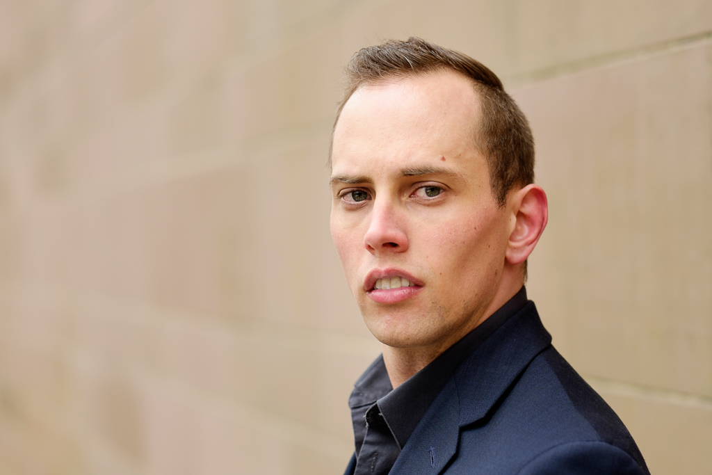 The author, a white male with brown hair, and navy suit jacket behind a beige brick wall.